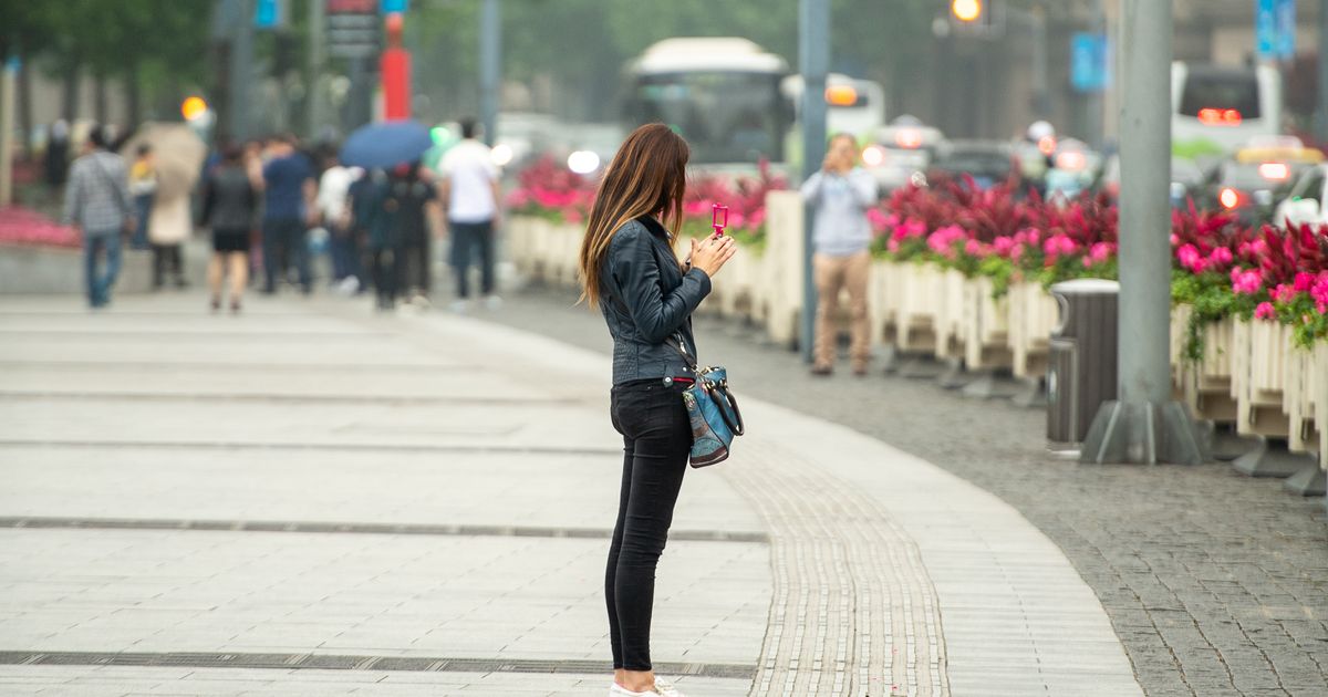 A traveler stands on a busy Shanghai sidewalk checking her smartphone beside flower planters and city traffic