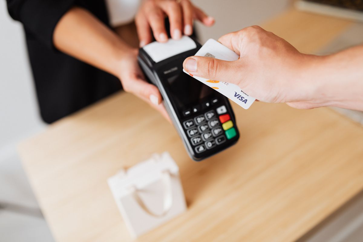 A hand tapping a Visa bank card on a black contactless card terminal at a shop counter