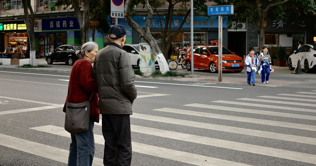 Two pedestrians waiting at a zebra crossing on a calm city street in Chengdu, China, with shops and trees lining the road