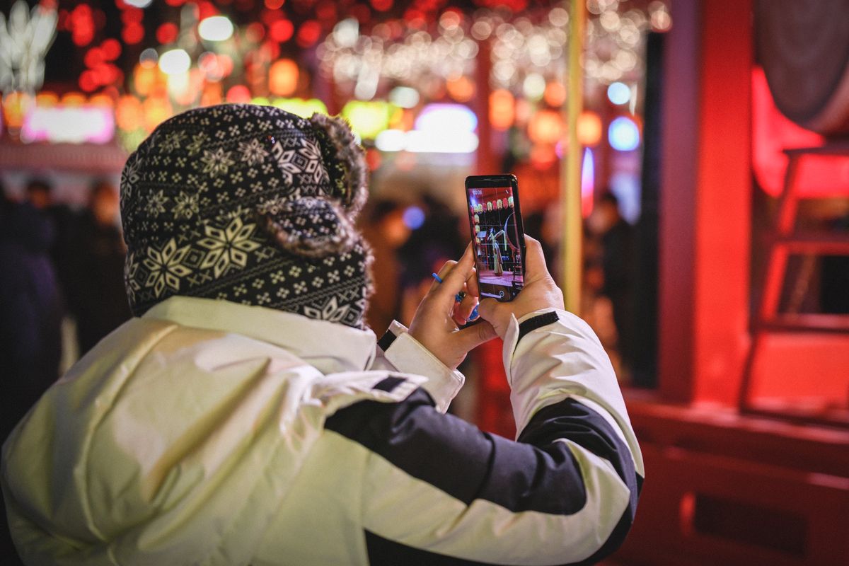 Traveler holding up a smartphone to photograph a lantern-lit Chinese night street, the scene visible on the phone screen