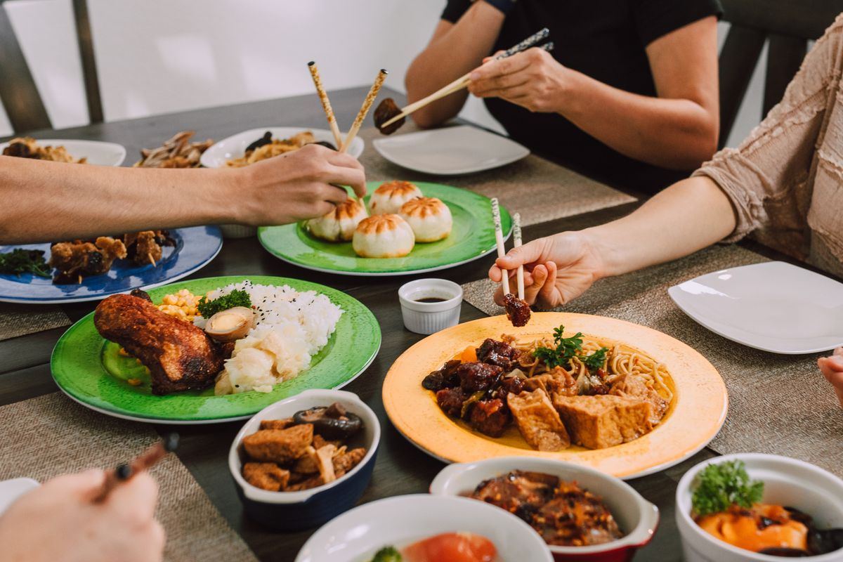 Several diners reaching across a table of shared dishes with chopsticks, including steamed buns and noodles